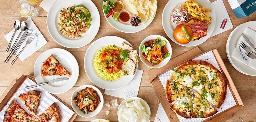 Assorted dishes sit ready to eat—pizzas, pasta, curry on rice, salad, burger with fries, poppadoms with dips—arranged on a wooden table with utensils, napkins, and takeout boxes. Text: HUGO'S.