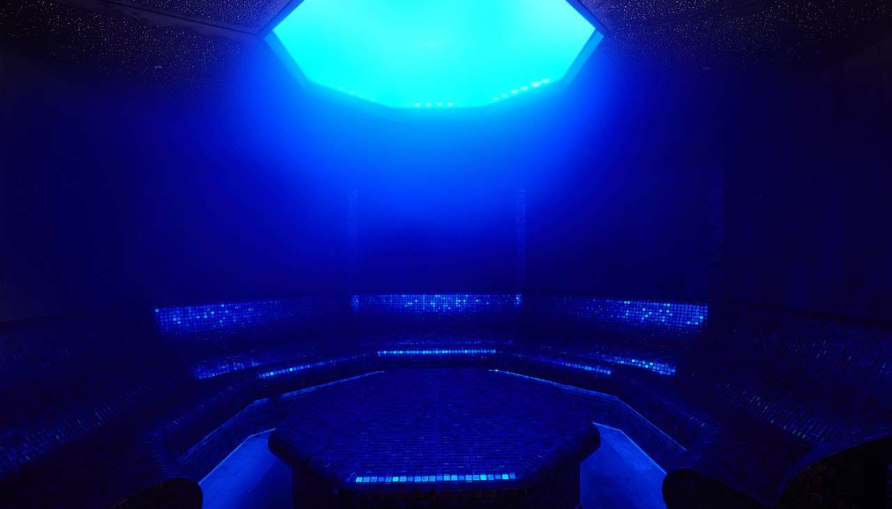 Blue lit steam room with sparkling black tiles and bench seating.