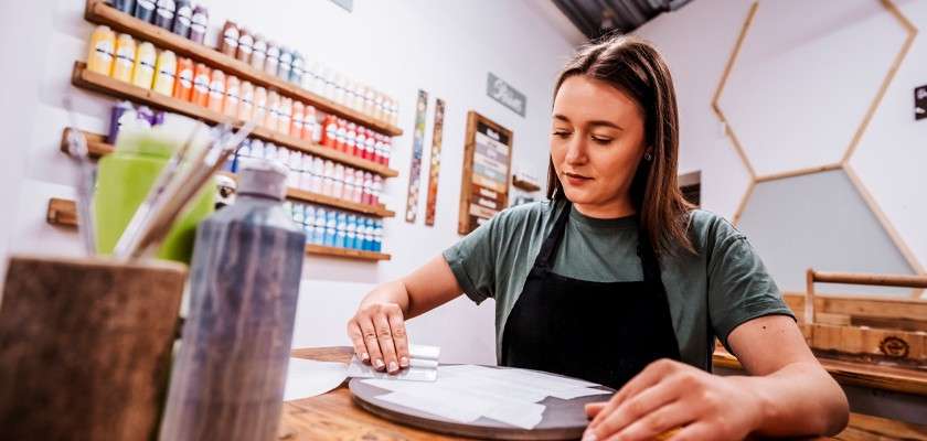 Person smooths a stencil on a round board, working at a wooden table in a craft studio; shelves of colorful paint bottles, brushes, and geometric wall decor surround them.