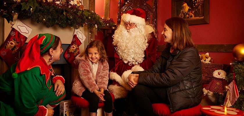 Child smiles while meeting Santa, chatting with an elf and a woman, in a cozy, warmly lit room decorated with garlands, stockings, presents, red walls, and festive ornaments.