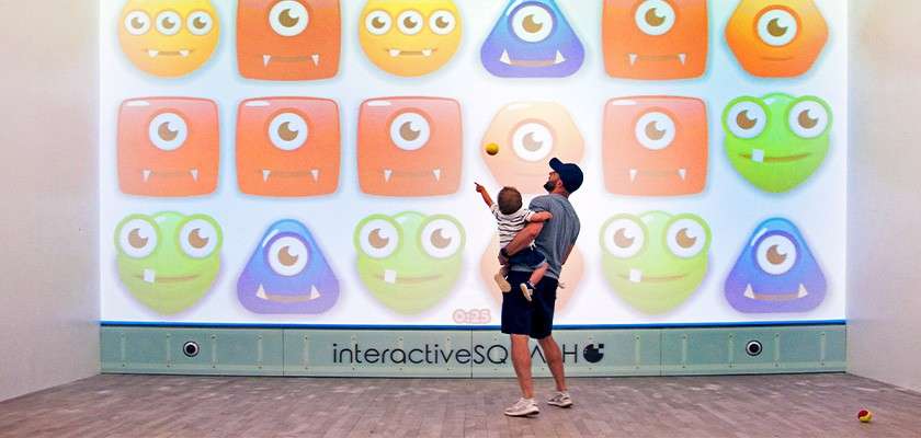 Adult holding a child throws a ball at a projected grid of cartoon monster faces, playing an interactive wall game in a room. Text: 0:25, interactiveSQU, H.