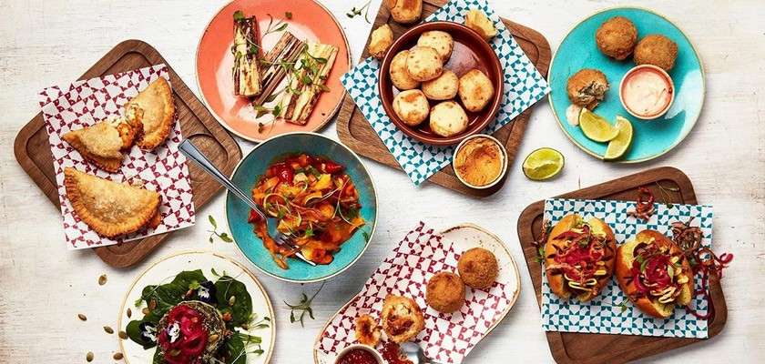 Assorted tapas-style dishes sit arranged: empanadas, croquettes, sliders, salad, grilled vegetables, and soups, with dipping sauces and lime wedges, on colorful plates and wooden boards across a white tabletop.