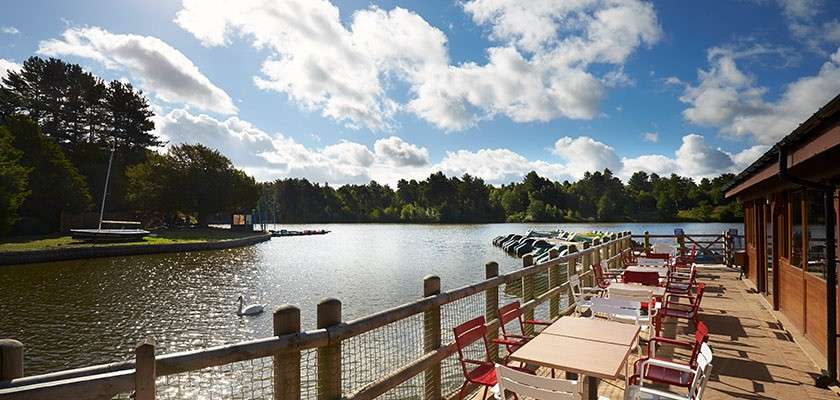 Outdoor lakeside patio overlooks calm water, empty tables and red chairs lined along railing. Pedal boats and small dock sit nearby, surrounded by trees under a bright, cloudy sky.