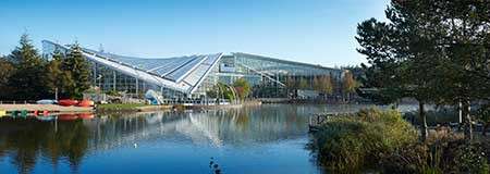 Glass-roofed leisure complex reflects across a calm lake, with docked red canoes nearby, surrounded by trees, reeds, and pathways under a clear blue sky.