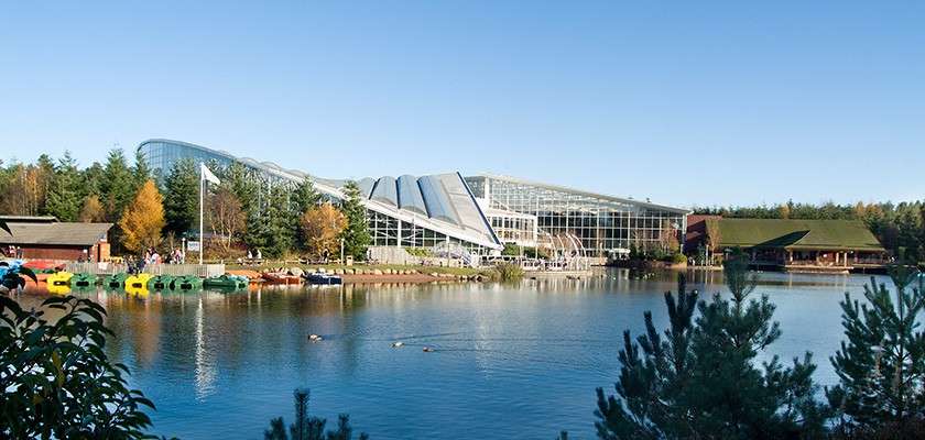 Glass-roofed leisure complex sits beside a calm lake, with paddle boats moored and a few people walking; surrounded by forested parkland, wooden lodges, and clear blue sky.