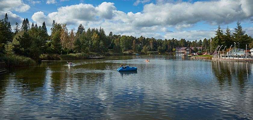 Small boats paddle across a calm lake, creating ripples. Forested banks, wooden walkways, and resort-style buildings frame the water under a partly cloudy sky.