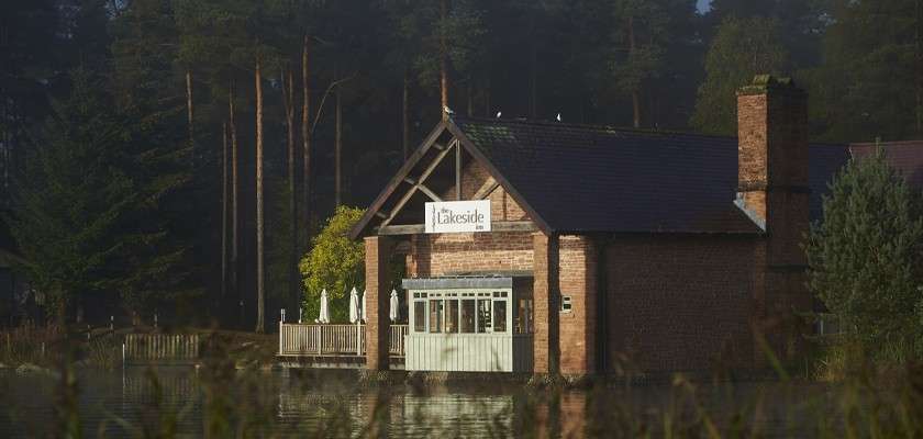 Brick lakeside café rests beside calm water; closed patio umbrellas stand on a wooden deck; tall pines surround the building. Visible sign text: The Lakeside.