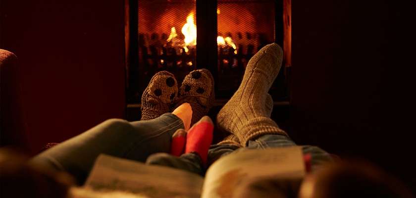 Two larger socked feet and a smaller red-socked pair rest, facing a lit fireplace; someone holds an open book in a dim, cozy room.