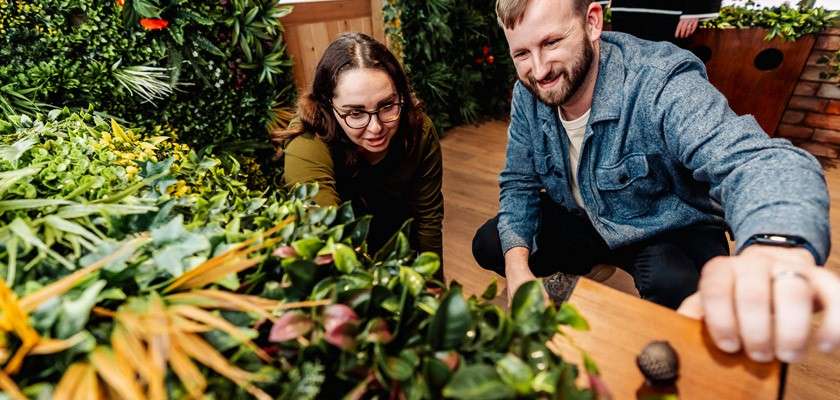 Two people examine leafy plants, one reaching toward a tabletop garden; surrounded by lush indoor greenery, wooden surfaces, and warm lighting in a plant-filled room.