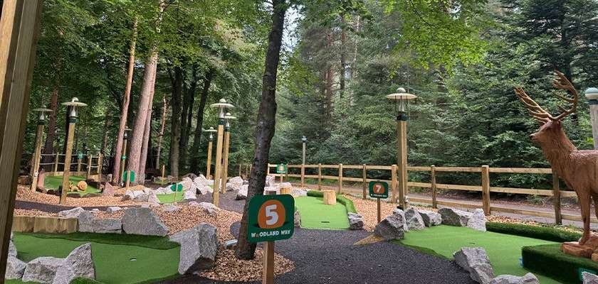 Mini-golf course—holes marked by signs—set in a wooded area with rocks, artificial turf, lamps, wooden fence, and a carved deer statue. Text: 5, WOODLAND WAY, 7.