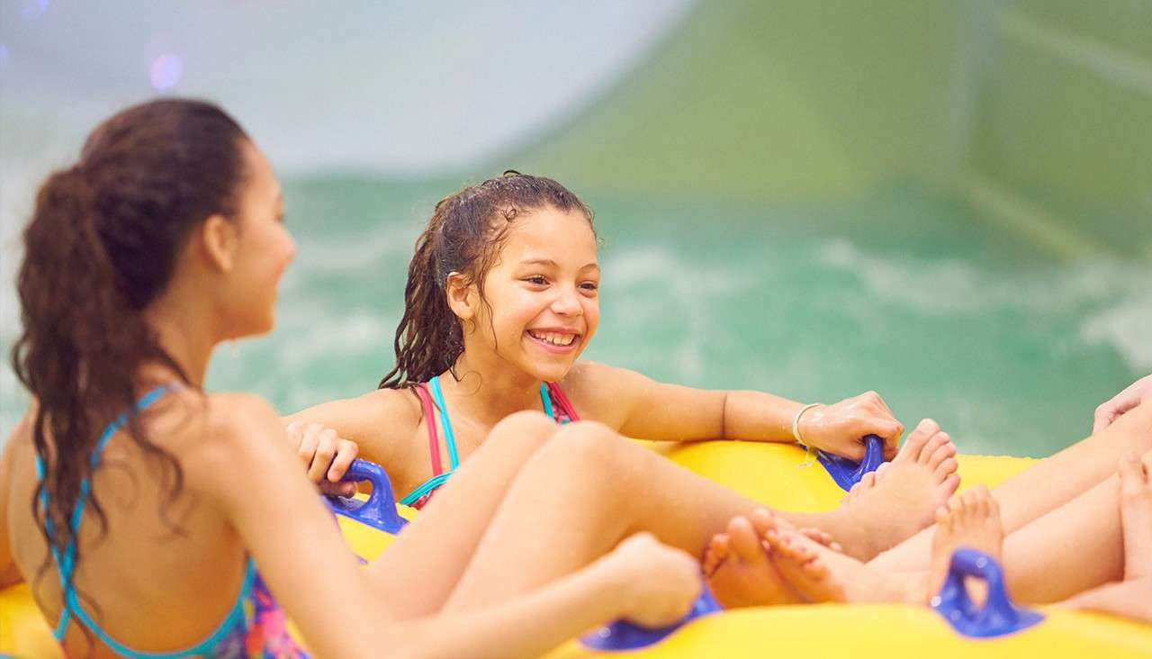 Two children smile and hold handles while riding a yellow inflatable raft, feet up, in choppy turquoise water at an indoor water park or pool slide.