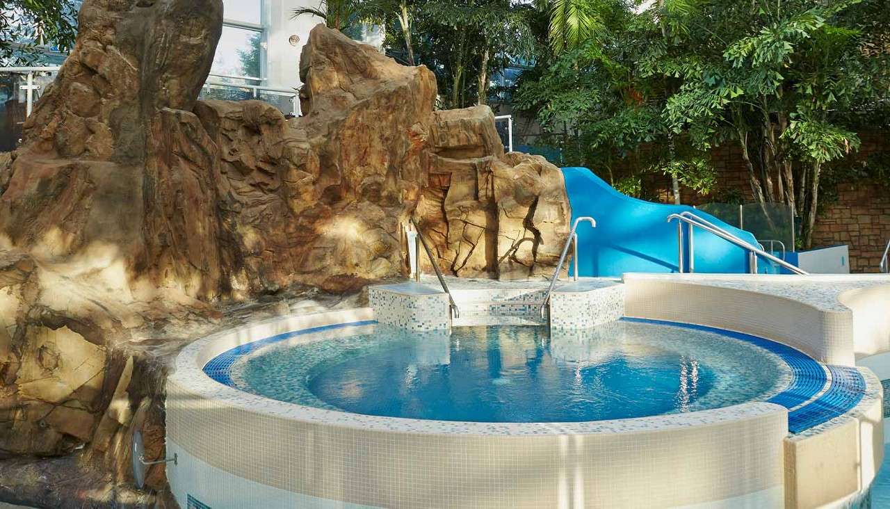 Circular hot tub bubbles with metal handrails and mosaic steps, beside faux rock wall and blue waterslide, set among tropical plants and glass windows in an indoor leisure complex.