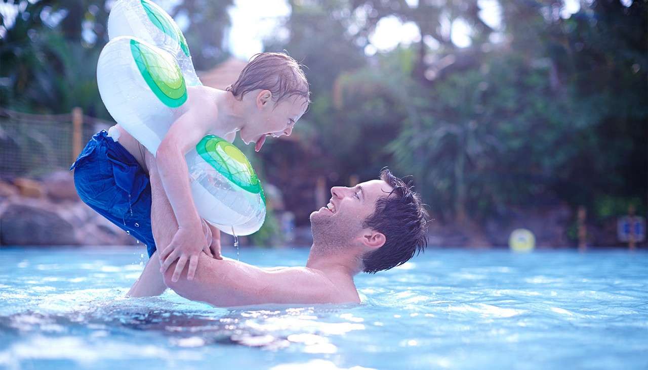 An adult lifts a laughing child wearing twin inflatable rings, water dripping, in a bright blue swimming pool, with tropical trees and sunlit bokeh in the background.