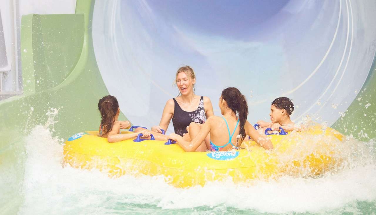 Four people ride a yellow inflatable raft, splashing out of a large enclosed waterslide, laughing and holding blue handles, surrounded by churning water inside a green-and-blue waterpark tube.