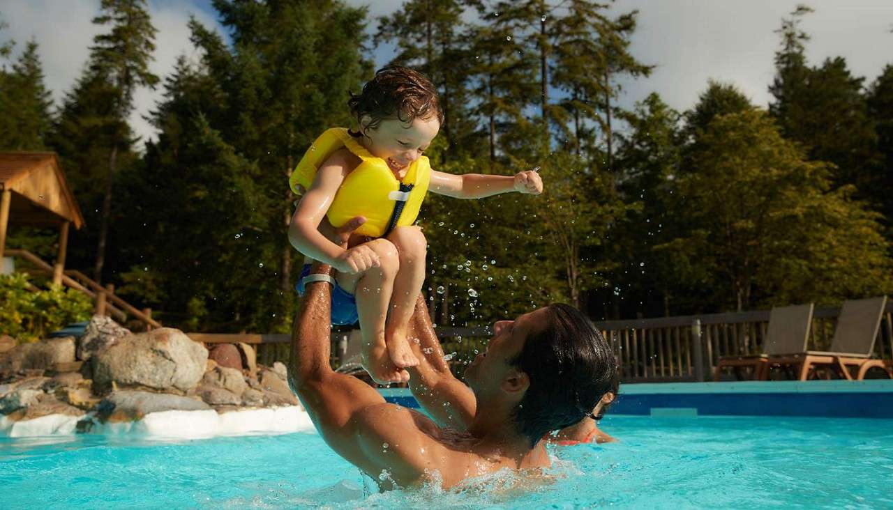Child in a yellow life jacket is lifted from the water by an adult; splashes rise in an outdoor swimming pool surrounded by trees, rocks, deck chairs, and sky.