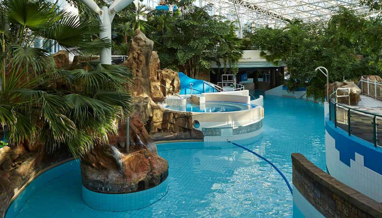 Indoor water-park pool curves as a lazy river flows; a small waterfall spills over faux rocks beside a blue slide. Tropical plants and walkways sit under a high glass-and-steel roof.