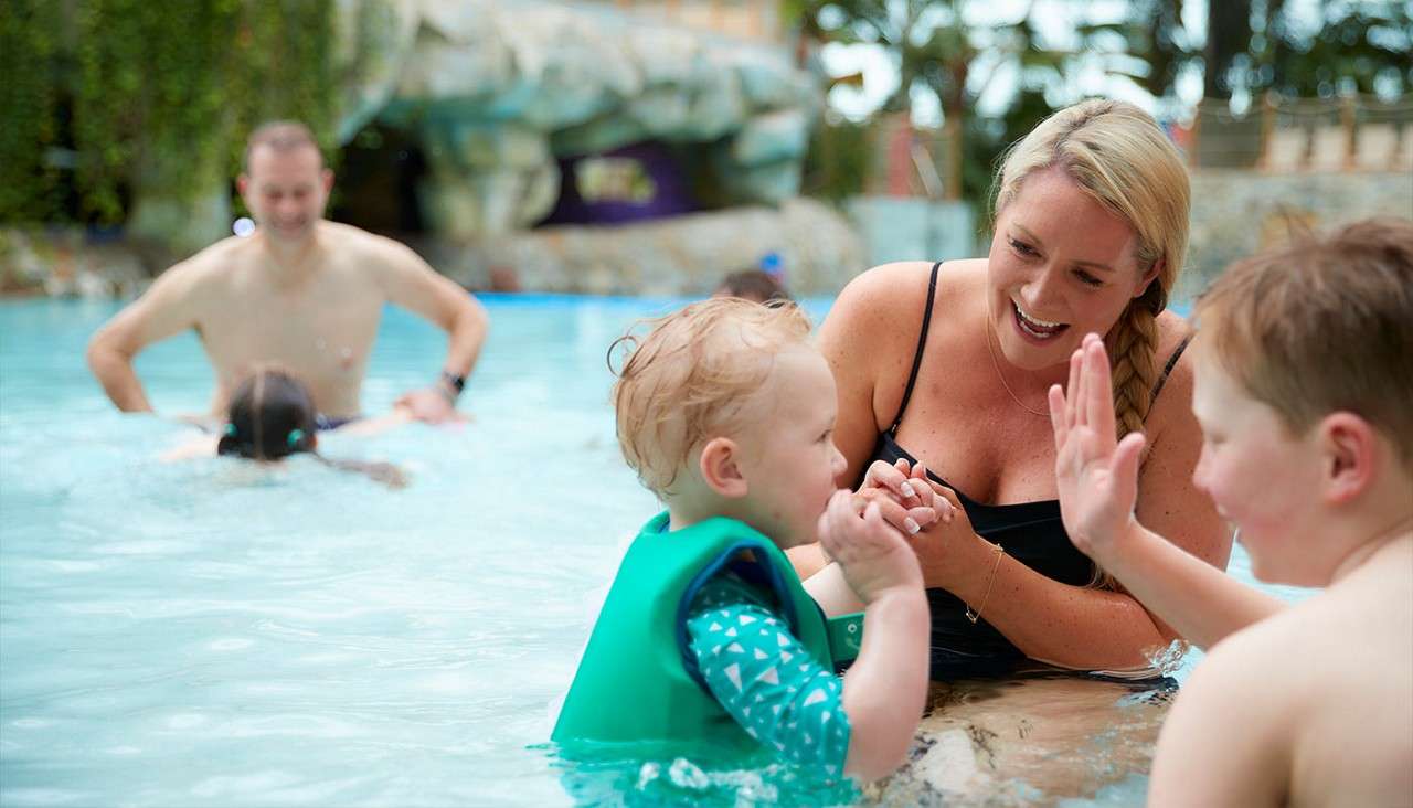 Toddler wearing a green flotation vest claps with a smiling woman and boy while wading; behind them, adults and a child swim in a shallow indoor pool.