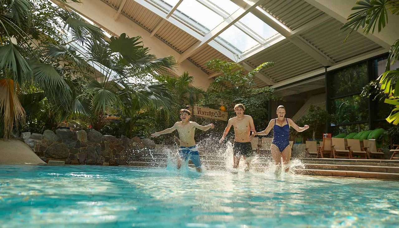 Three people run into a shallow pool, splashing water, inside an indoor tropical waterpark with palm plants, skylights, and lounge chairs. Text visible on a sign: “Venture Bay”.