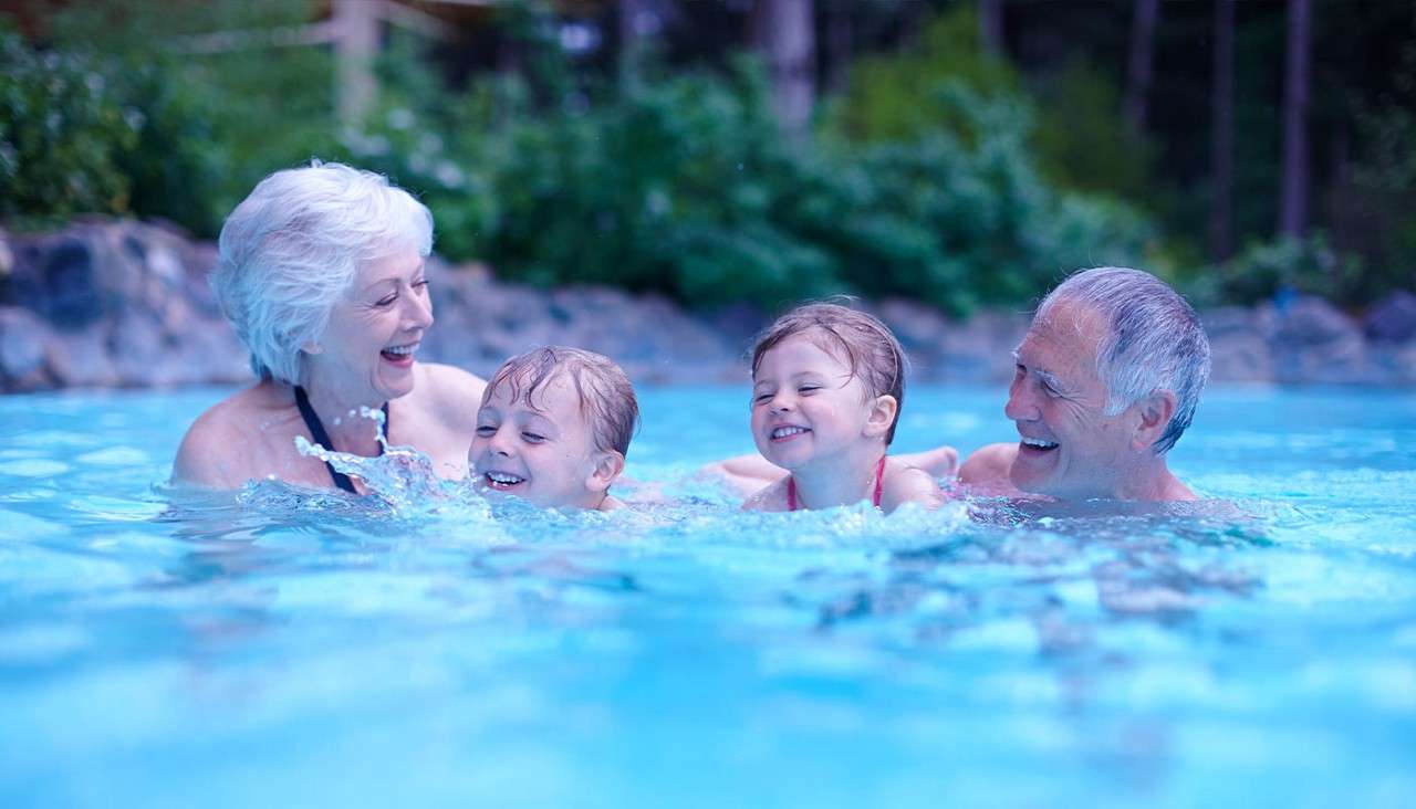 Two older adults splash and support two laughing children, swimming together in a bright blue pool; trees and rocks surround the outdoor setting.