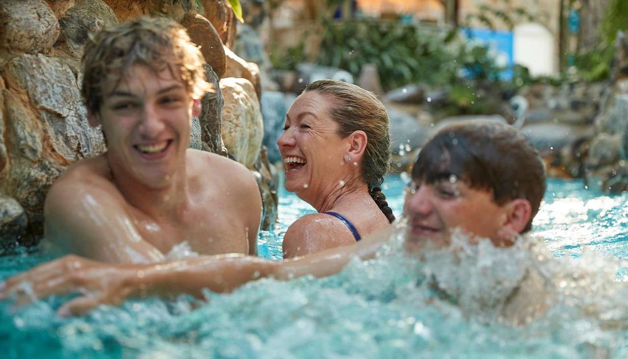 Three swimmers laugh and splash, bobbing in turquoise water near rock walls, in an indoor pool area with plants and warm light.