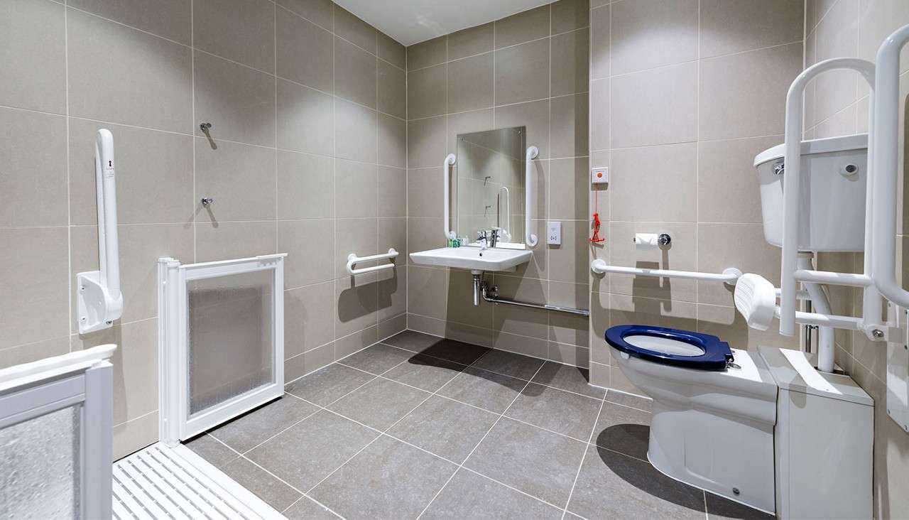 Accessible toilet and sink assist users, surrounded by grab bars, raised seat, and emergency pull cord, in a spacious, beige-tiled bathroom with non-slip floor and walk-in shower area. Text: 1.