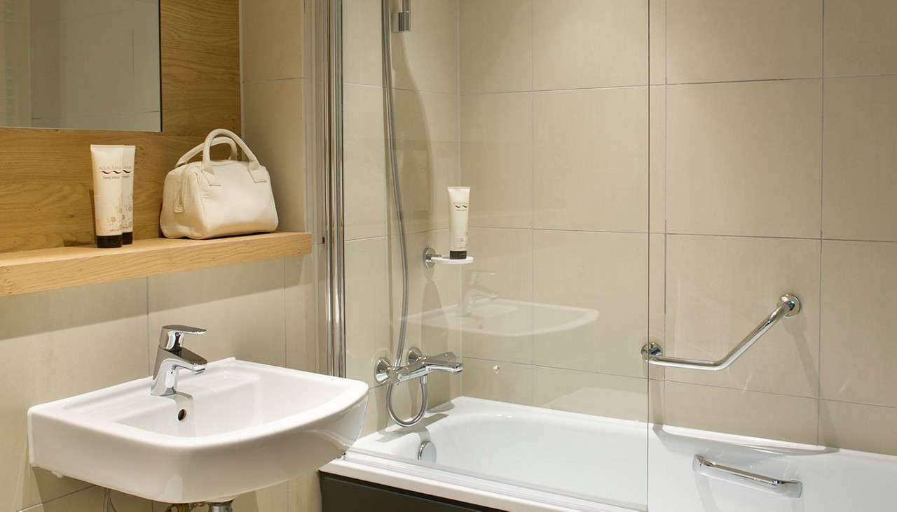 Bathtub with glass screen stands against tiled wall; handheld shower hangs. Nearby, a wall-mounted sink sits below a wooden shelf holding toiletries and a white bag, with chrome grab bars installed.