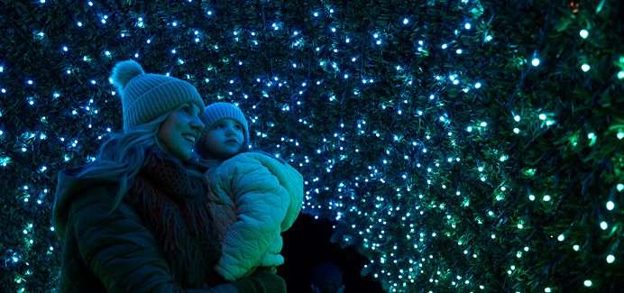 Adult holding a bundled child gazes upward, standing inside a tunnel of blue-green string lights, both wearing knit beanies and winter coats in a nighttime display.