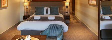 Bed sits neatly made with folded towels on top, centered under twin lamps. Surroundings: carpeted hotel room with armchair and table, framed art, mirror, and corridor leading toward bathroom area.