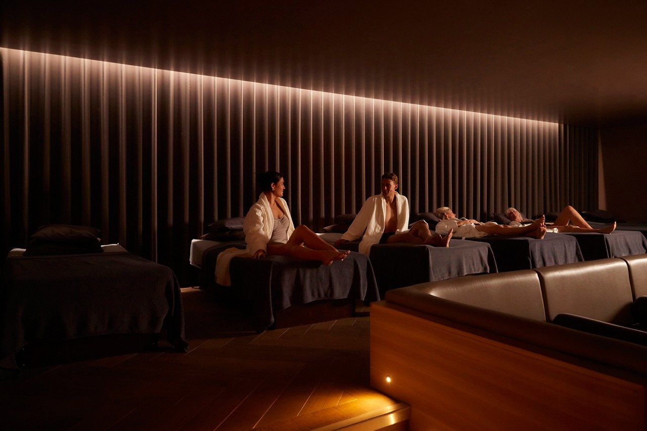 Spa guests lounge and chat on padded daybeds while others recline resting. Warm, low lighting and ribbed wall panels create a calm, dim relaxation-room ambiance.