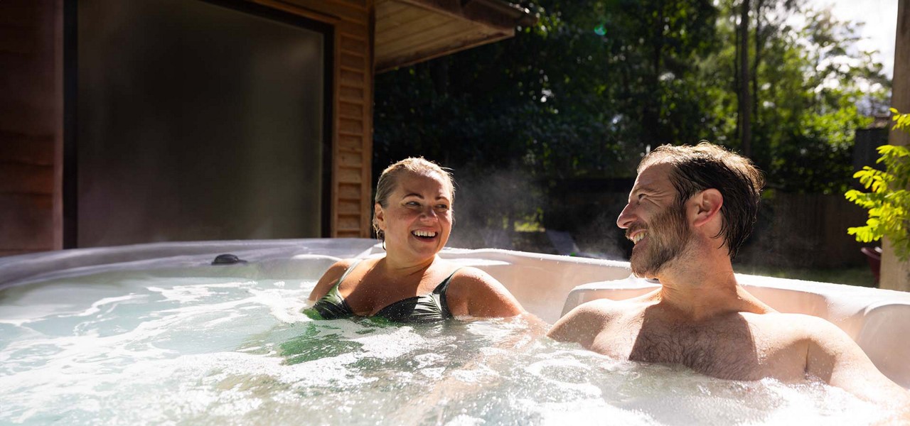 Two adults relax and chat in a bubbling hot tub, smiling at each other, as steam rises in sunlight; beside a wooden building, surrounded by trees in a backyard setting.