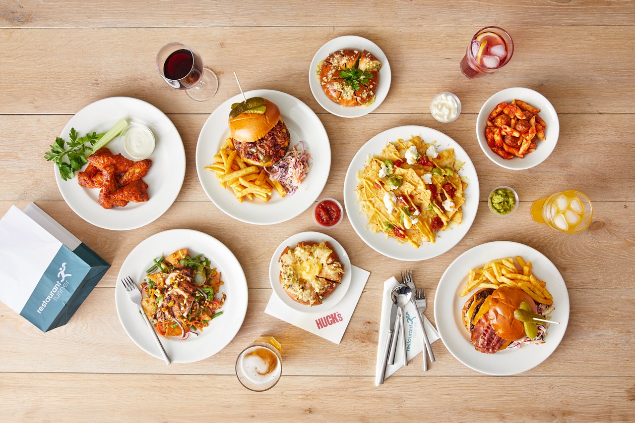 Assorted plated meals—burgers with fries, nachos, wings, salad, pasta, pretzel bites—sit arranged on a wooden table with beer, wine, iced drinks, utensils, and branded items. Text: restaurant runner; HUCK's.