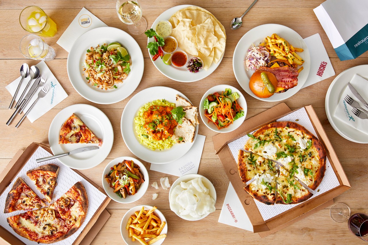 Assorted dishes arranged for sharing on a wooden table: pizzas, curry with rice, noodles, burger with fries, salads, dips, and drinks. Text: “PUNCH”, “HUCK’S”, “RAJINDA PRADESH”, “restaurant”, “PIZZARRIO”.