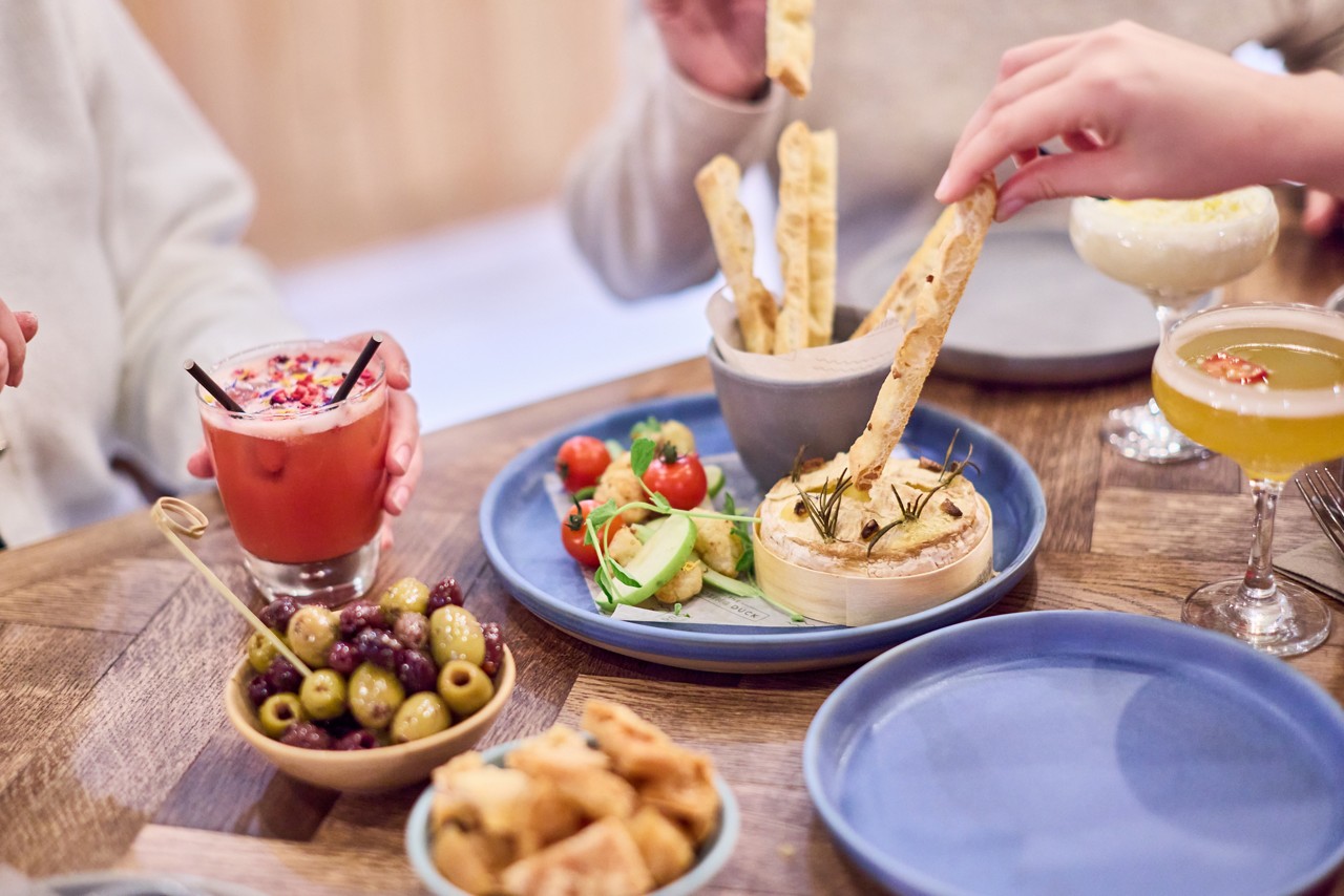 Hands dip breadsticks into baked cheese while others hold cocktails, on a wooden table set with olives, cherry tomatoes, cucumber, fried bites, and blue plates in a casual dining setting.