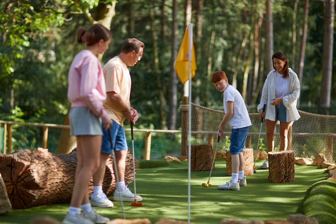 A boy putts a mini-golf ball while three companions watch and prepare, on an outdoor course with faux logs, netted barriers, and trees in the background.