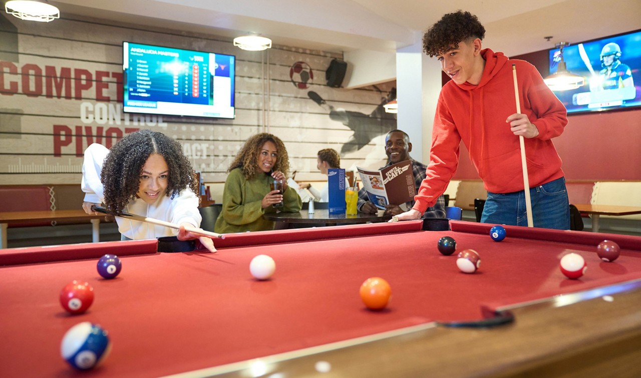 Two young people play pool, leaning over a red table; friends watch with drinks in a sports bar with TVs. Visible text: COMPETE; DRINKS; ANDALUCIA MASTERS; PROFESSIONAL LEAGUE.