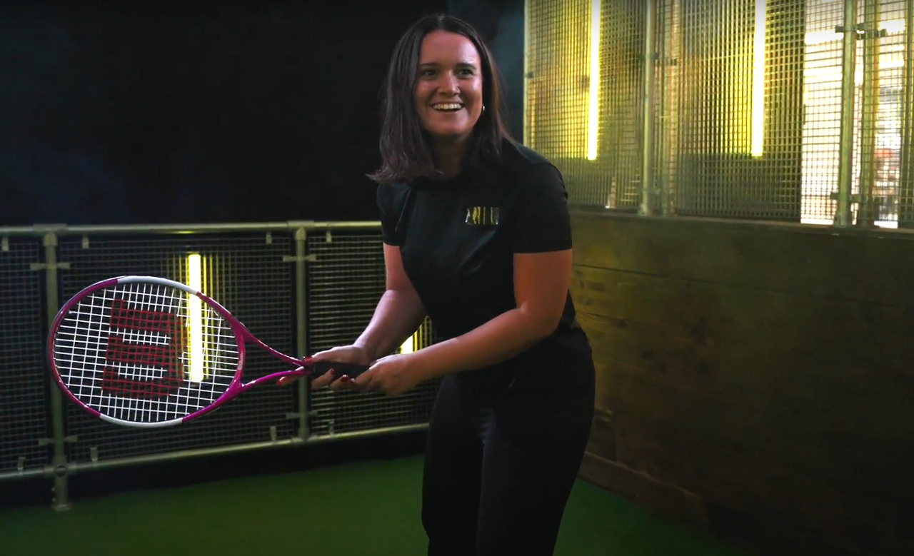 Woman holding a tennis racket inside The Bat Cage.