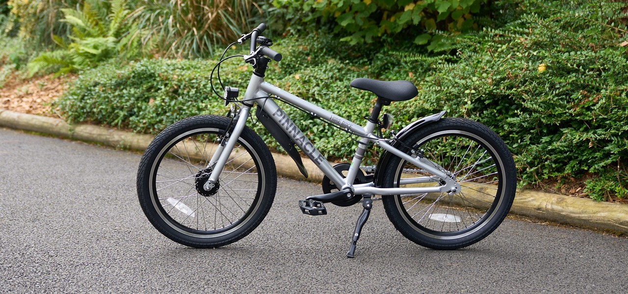 Children’s silver bicycle stands on a kickstand on a paved path; front and rear fenders, reflectors, and thick tires visible; frame text reads “PINNACLE” and “HIRE BIKE”; bushes and curb behind.