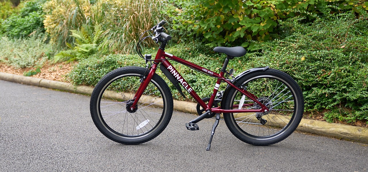 Red bicycle stands on its kickstand on a paved road, near dense greenery and a curb. Text on frame: PINNACLE, HIRE BIKE.