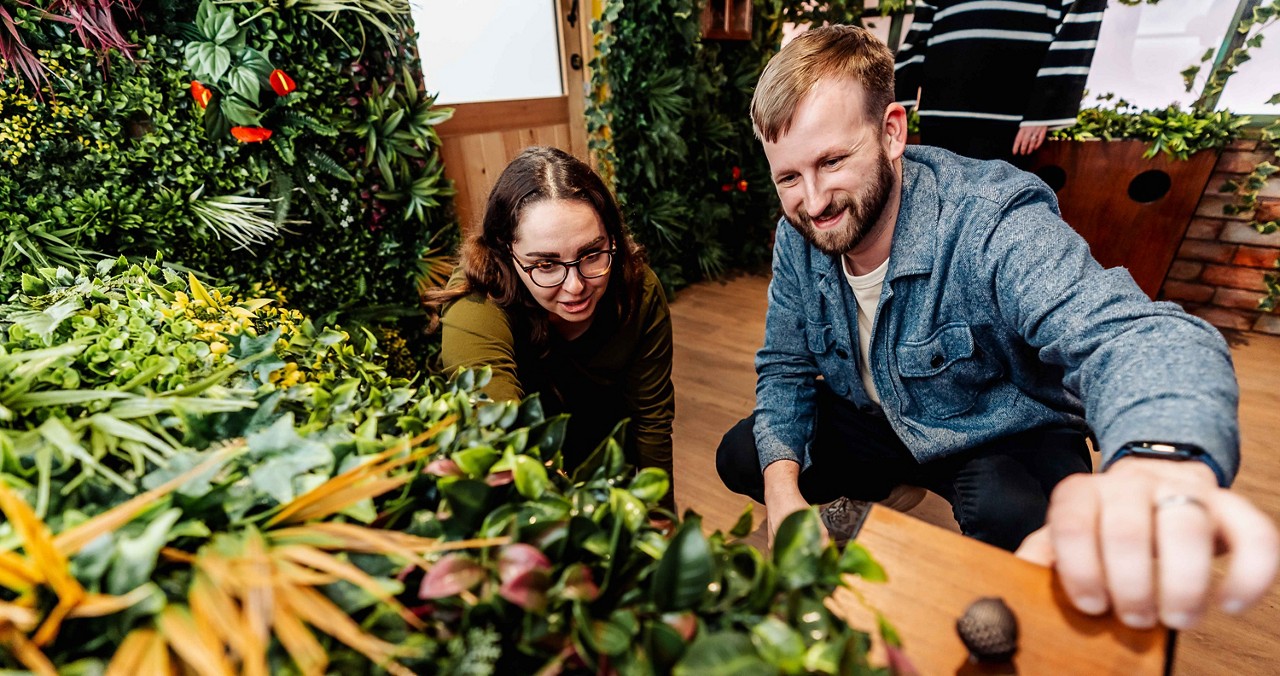 Two people kneel and examine leafy plants, smiling and reaching toward a planter. They are indoors, surrounded by dense greenery on walls and shelves, with wooden flooring and natural light.
