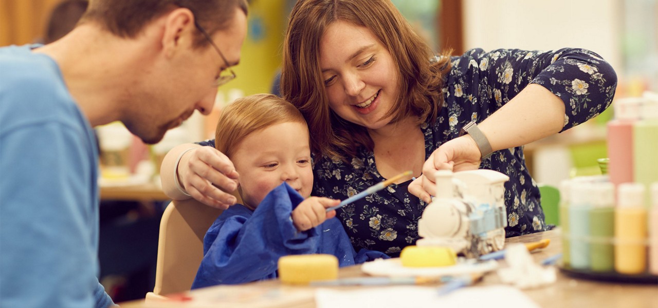 Child with paintbrush paints a ceramic train as two adults guide and smile, at a craft table in a bright studio with bottles of paint, sponges, and scattered tools.