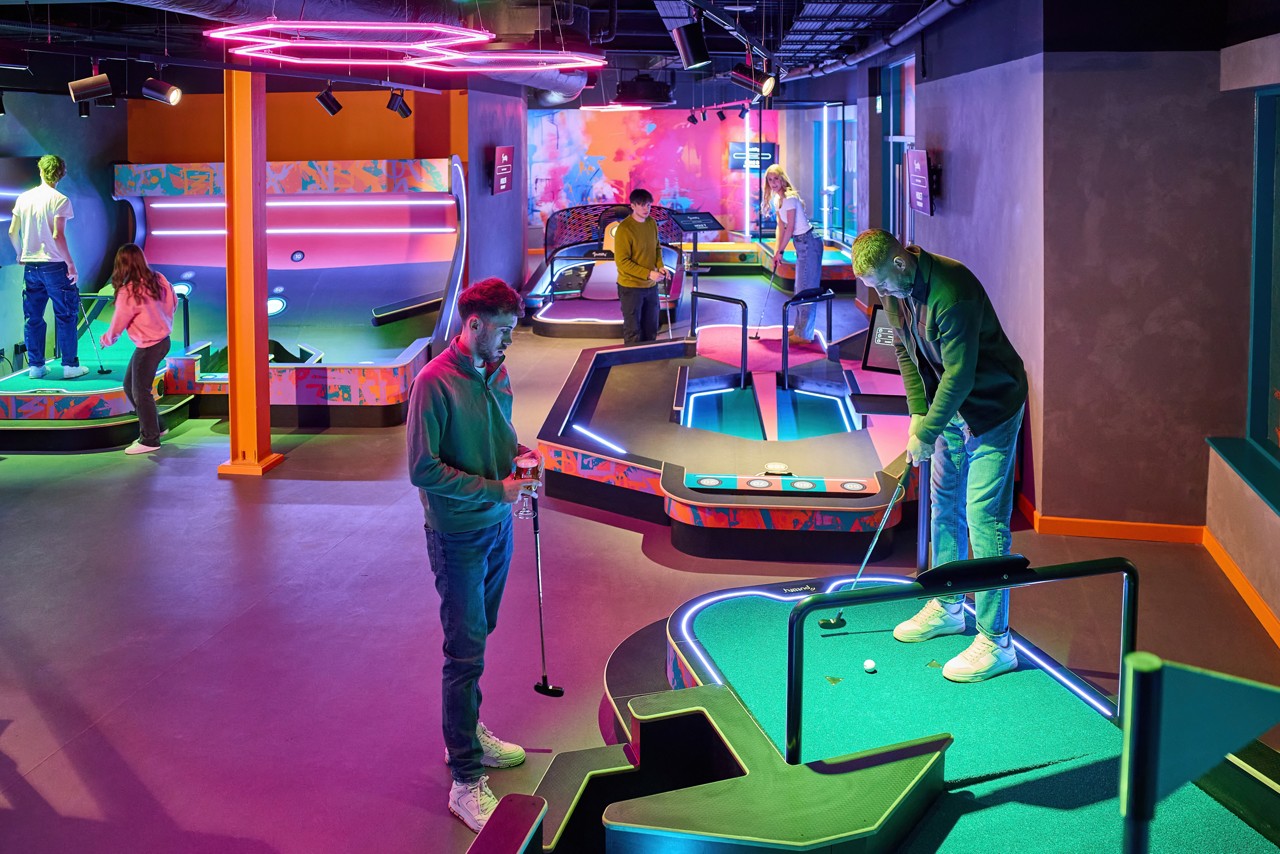 Mini-golfers putt on glowing, obstacle-filled lanes in a neon-lit indoor arcade; multiple groups play simultaneously among colorful ramps, rails, and futuristic lighting fixtures.