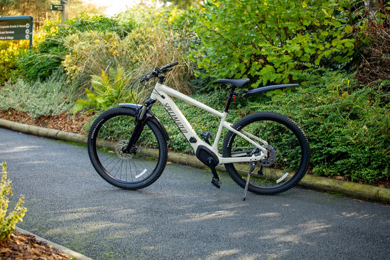 Electric mountain bike stands parked on its kickstand, facing left, on a paved garden path amid bushes and trees; front suspension and fenders visible. Text on frame: "SPECIALIZED".