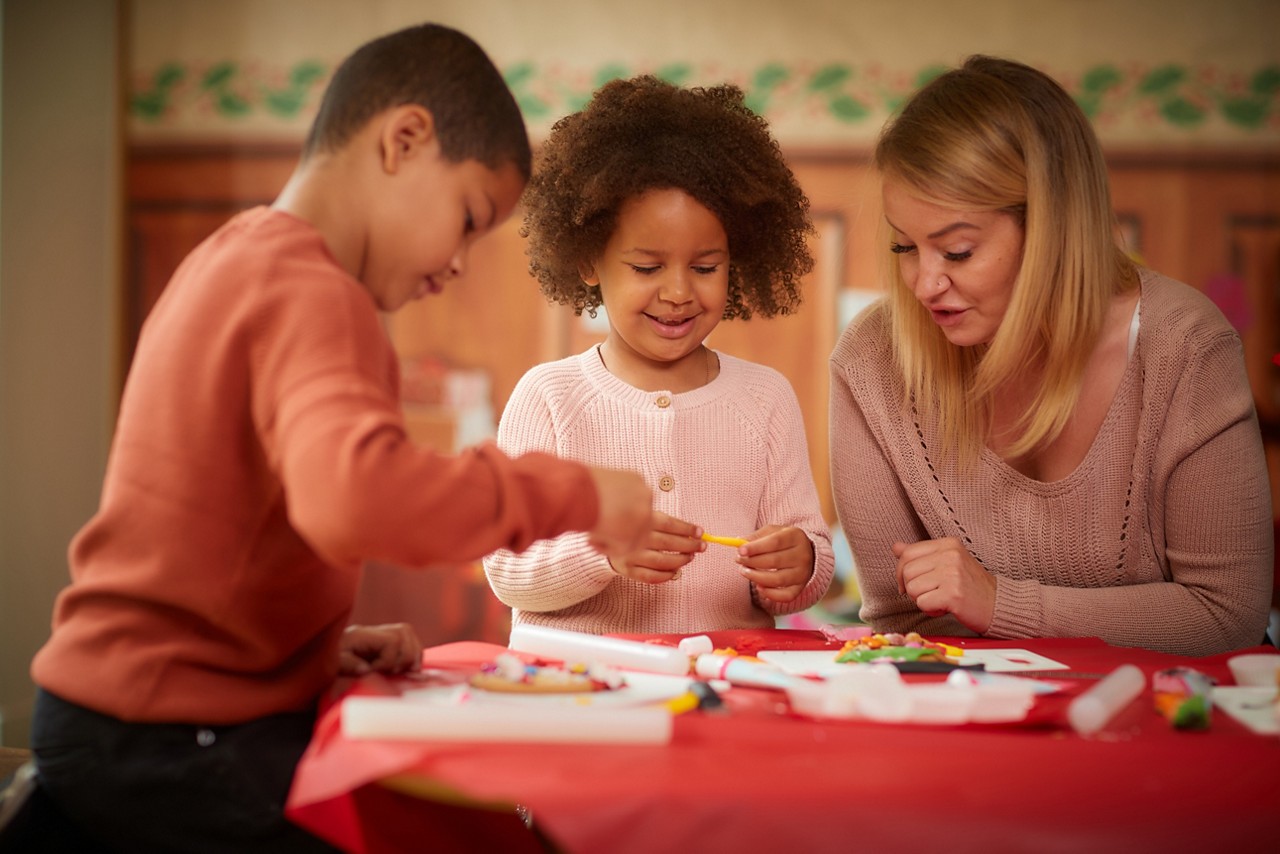 Children and an adult assemble crafts, handling markers and small objects at a red-covered table in a lit classroom with wooden cabinets, a holiday-themed wall border, and scattered art supplies.