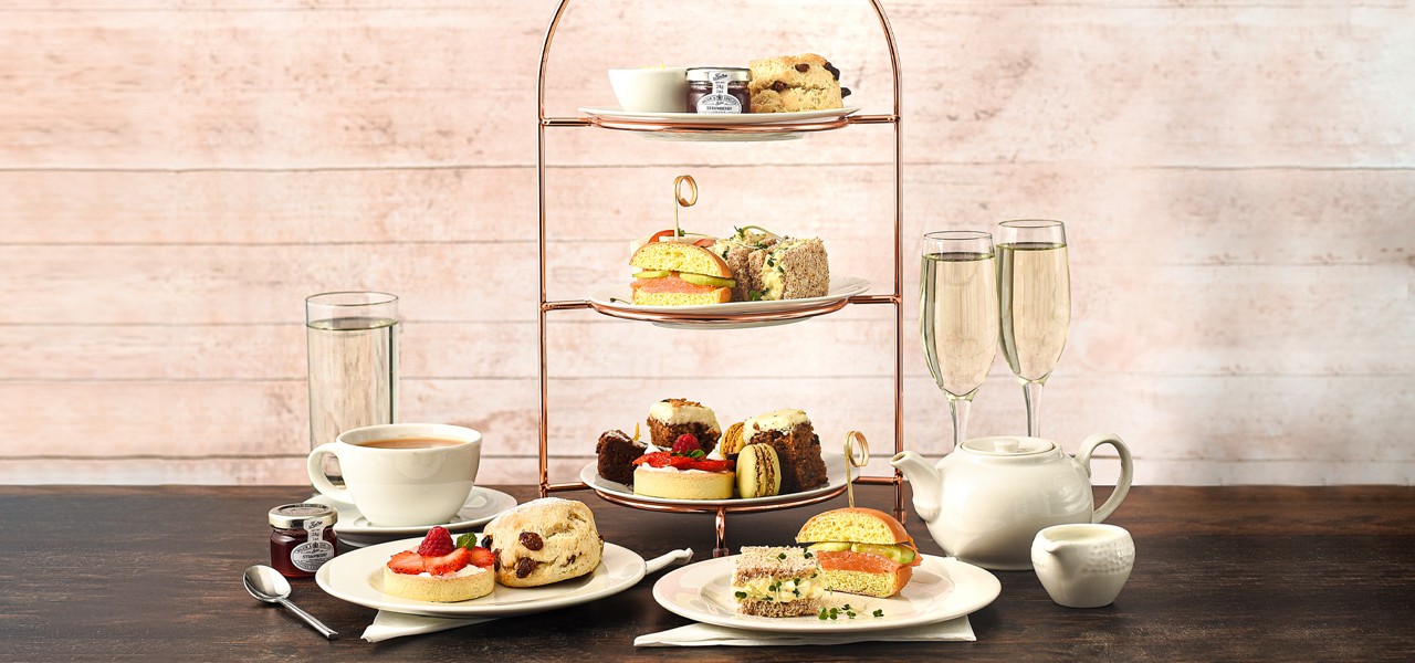 Three-tier afternoon tea stand presents scones with cream and jam, assorted pastries, and sandwiches; beside it rest a white teapot and cup on a wooden table against a dark backdrop.