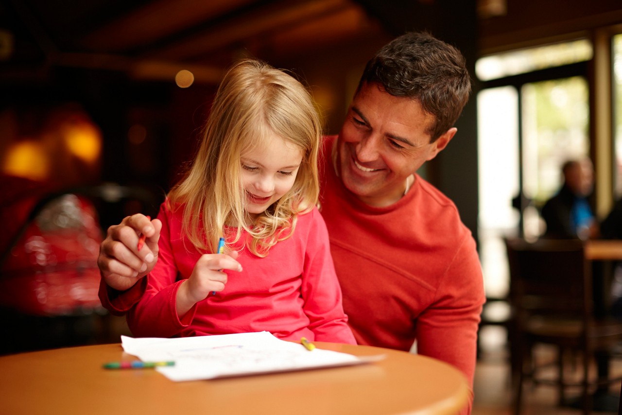 A little girl sat on her Dad's knee colouring in a picture at the table. 