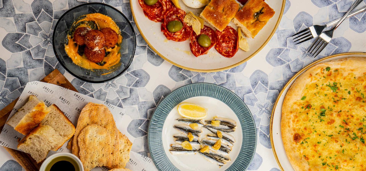 Assorted Italian dishes sit arranged on a patterned tabletop: garlic pizza, antipasto with salami, olives, focaccia, anchovies with lemon, arancini, and bread with oil. Text: AMALFI AMALFI AMALFI AMALFI AMALFI AMALFI AMALFI AMALFI.