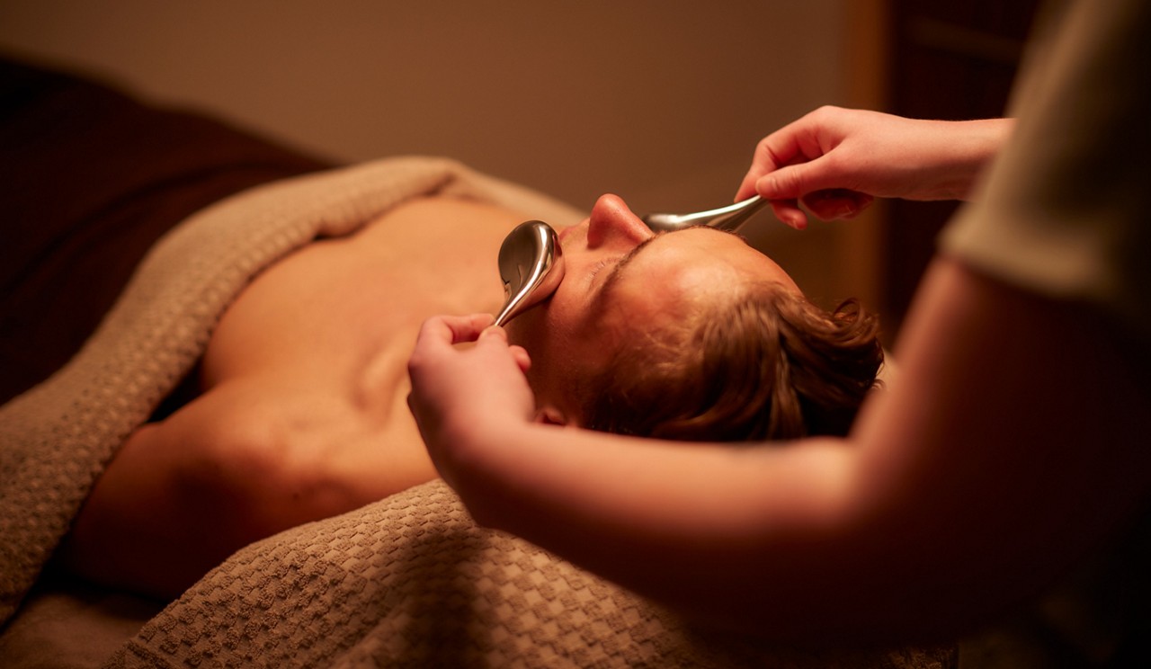 Person’s face—eyes closed—receives a facial massage as two metal spoons glide along cheeks and nose; body lies under a textured towel on a massage table in warm, dim spa lighting.
