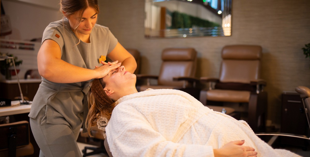 Esthetician massages a client’s forehead; the client reclines in a white robe on a salon chair. Background shows cushioned pedicure chairs, mirror, and nail station. Visible text: OPI.