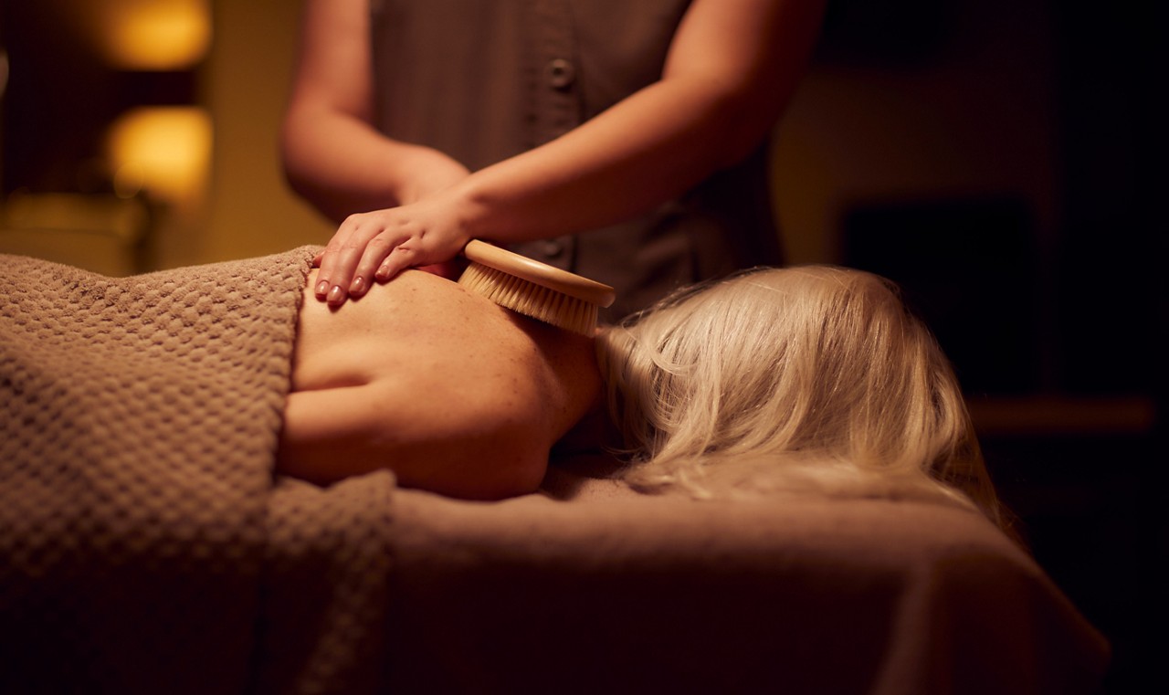 Client lies face down on a massage table; therapist uses a wooden-bristled dry brush on the shoulder; warm, dim spa room with a textured blanket covering the body.