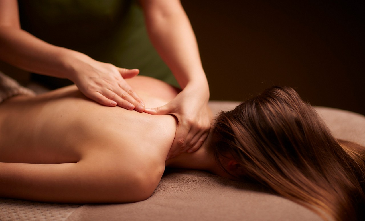 Massage therapist presses and kneads a woman's bare upper back and neck, applying pressure with both hands; client lies face down on a padded table in a dim, spa-like room.